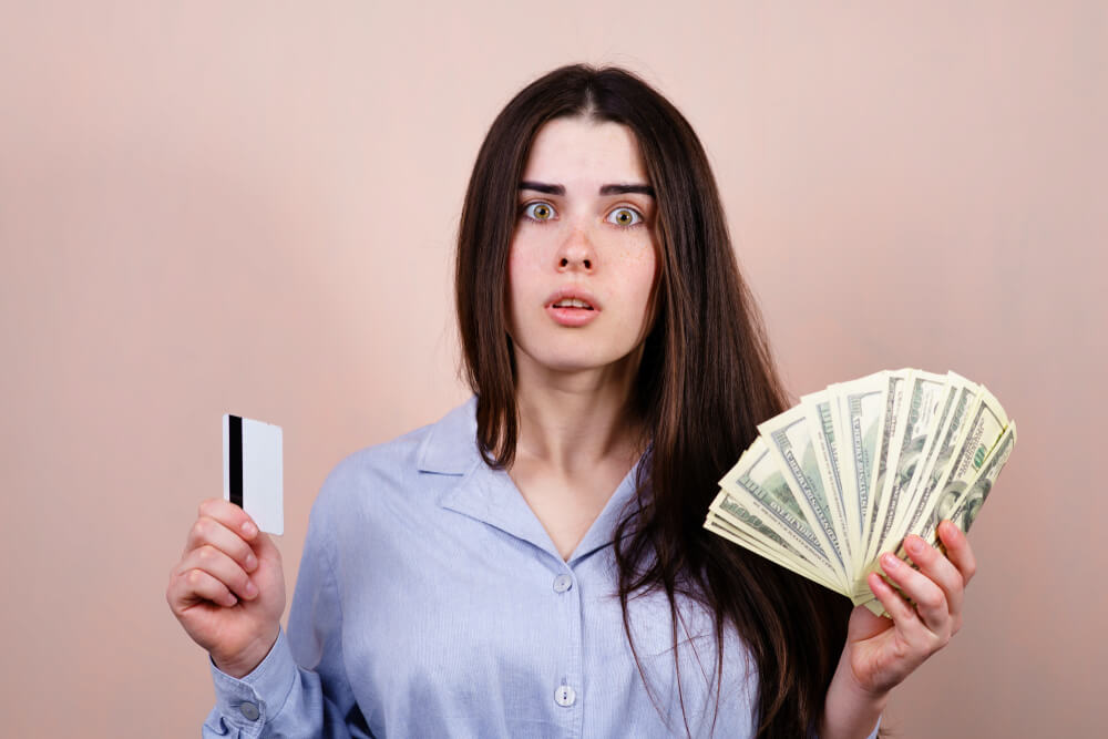 Electronic Money vs Cash. Young Pensive Woman With Credit Card and Dollar Bills.