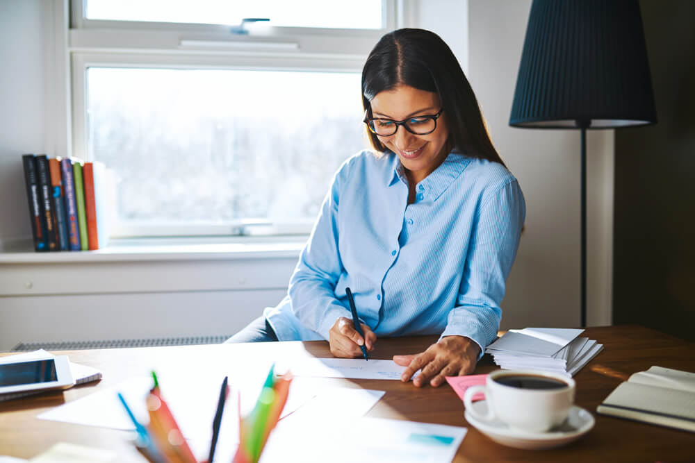 Beautiful Young Self-Employed Woman Wearing Glasses and Blue Shirt at Desk Writing Checks Next to Cup of Coffee and Booklets