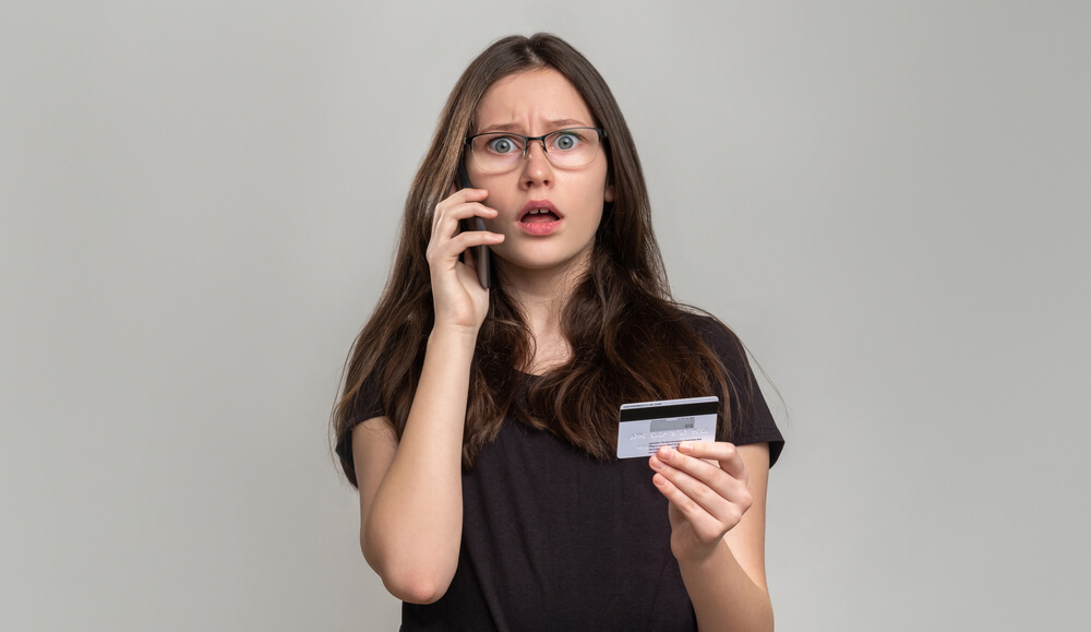 Shocked Woman Checking Credit Card Balance on Phone Isolated on Gray.